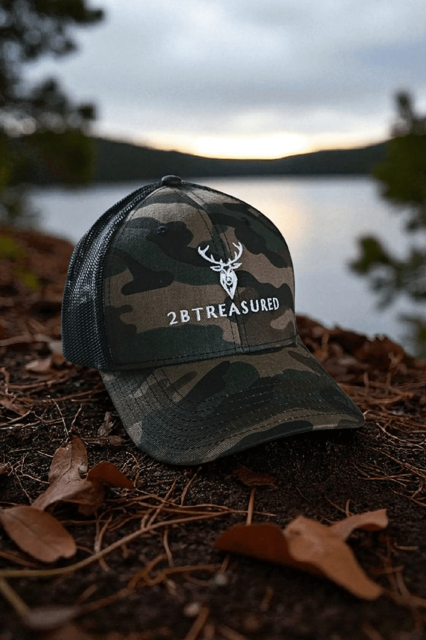 Embroidered Camouflage Trucker Hat front view from 2BTreasured resting of forest floor with lake in background