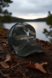 Embroidered Camouflage Trucker Hat front view from 2BTreasured resting of forest floor with lake in background