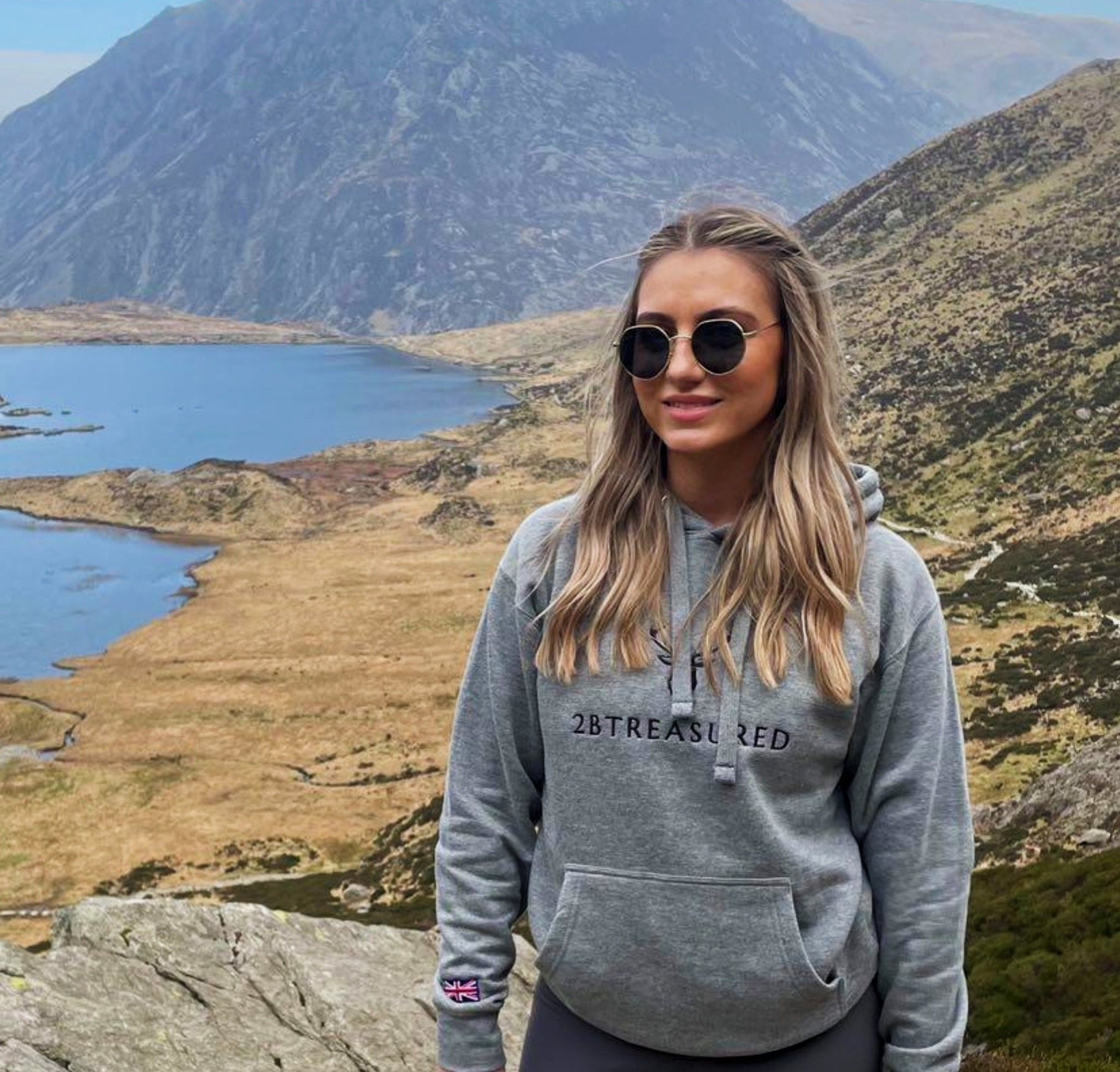 Female wearing the Embroidered Lomond Hoodie with British Flag in grey standing on a hill with mountains in the background 