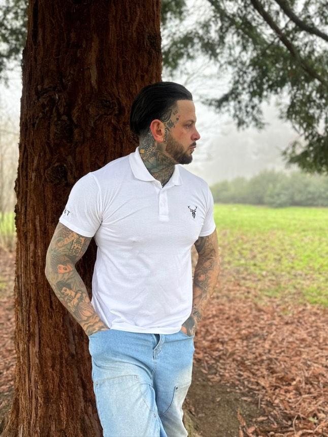 Male with tattos wearing the Embroidered Premium Hillside Polo Shirt in white from 2BTreasured next to a tree